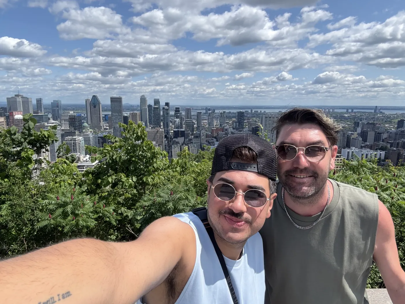 Christopher and Jake in sunglasses at a hilltop lookout, a city skyline and river spread out behind them.