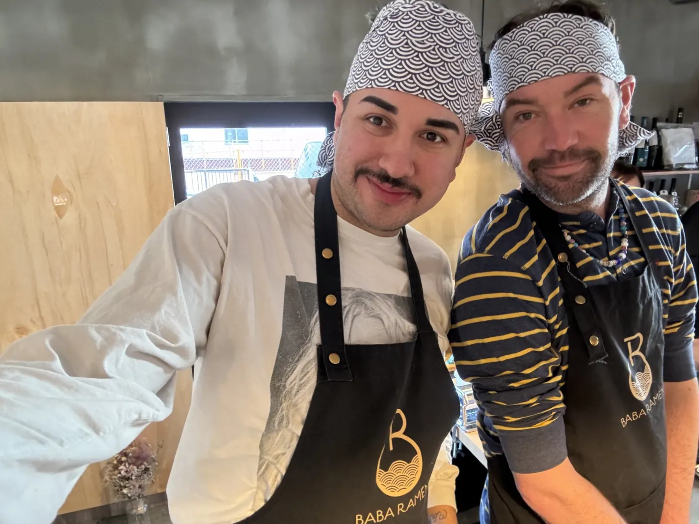 Christopher and Jake behind the counter at Baba Ramen in Japan, both in wave-print bandanas and shop aprons.