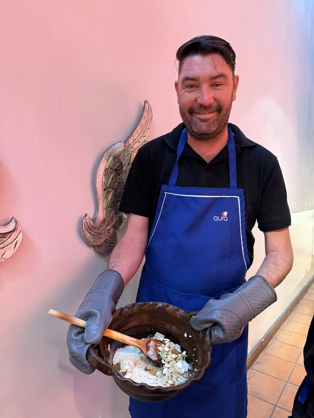 Christopher Fairman in a blue apron and oven mitts, grinning as he holds a clay bowl of spices at a cooking class in Mexico City.