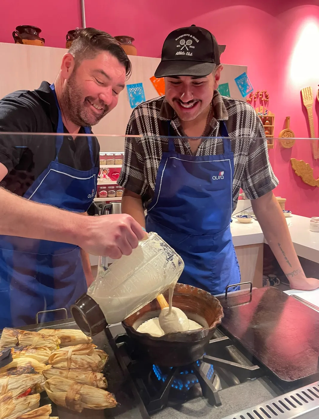 Christopher laughing as he pours masa batter into a clay pot over a flame, a tray of tamales beside him, in a bright pink Mexico City kitchen.