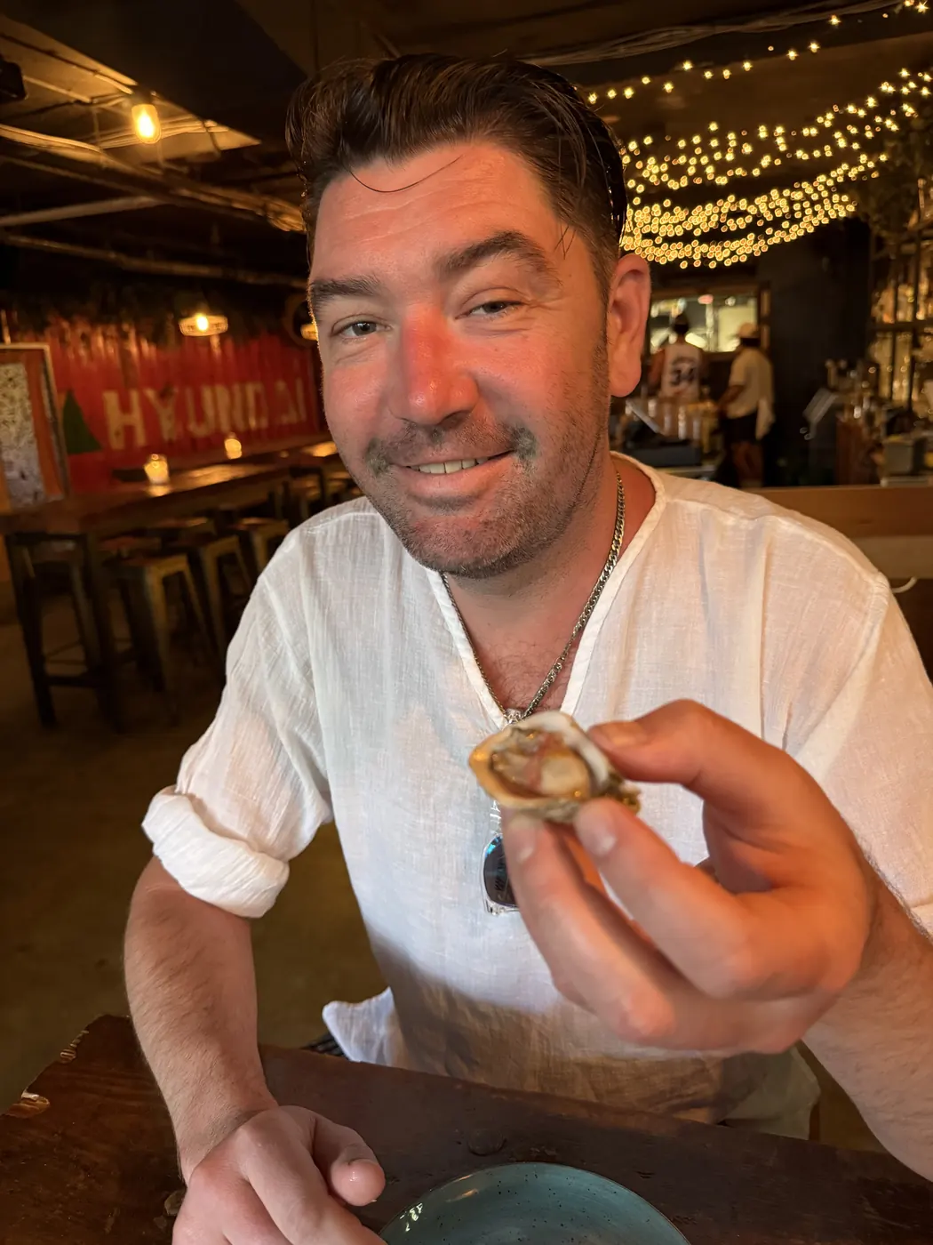Christopher holding up a shucked oyster at a string-lit bar, smiling at the camera.