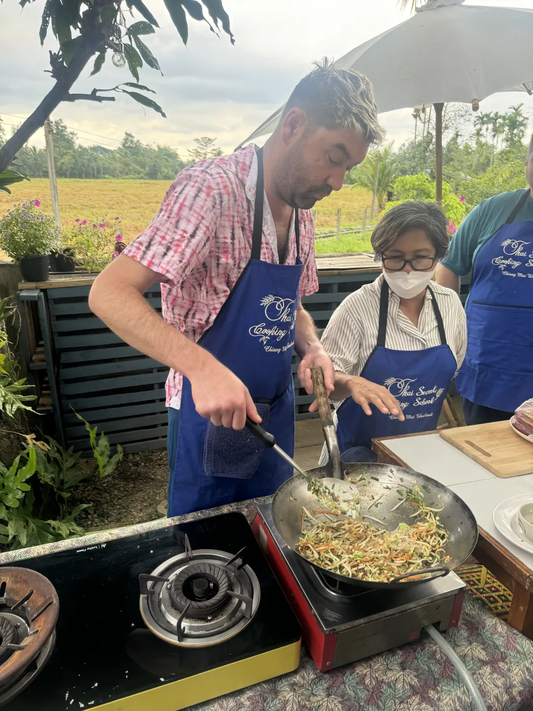 Christopher tossing vegetables in a wok over a gas burner at an open-air cooking school beside a rice field in Chiang Mai, Thailand.